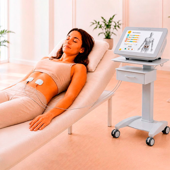 Eine Frau ruht entspannt auf einer Behandlungsliege. Elektroden sind am Bauch angebracht, daneben steht das Frequency First Gerät